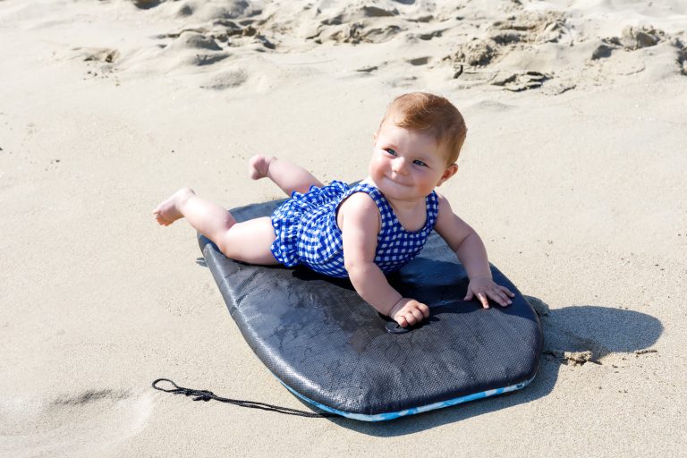 Das Foto zeigt ein Baby im Badeanzug am Strand auf einem Surfbrett.